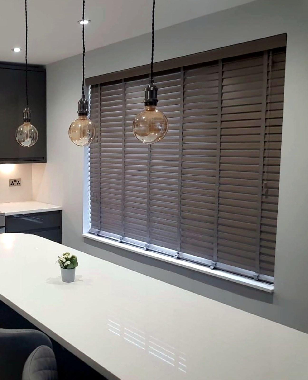 Grey wood venetian blind fitted to a kitchen window, with pendant lights over a white worktop