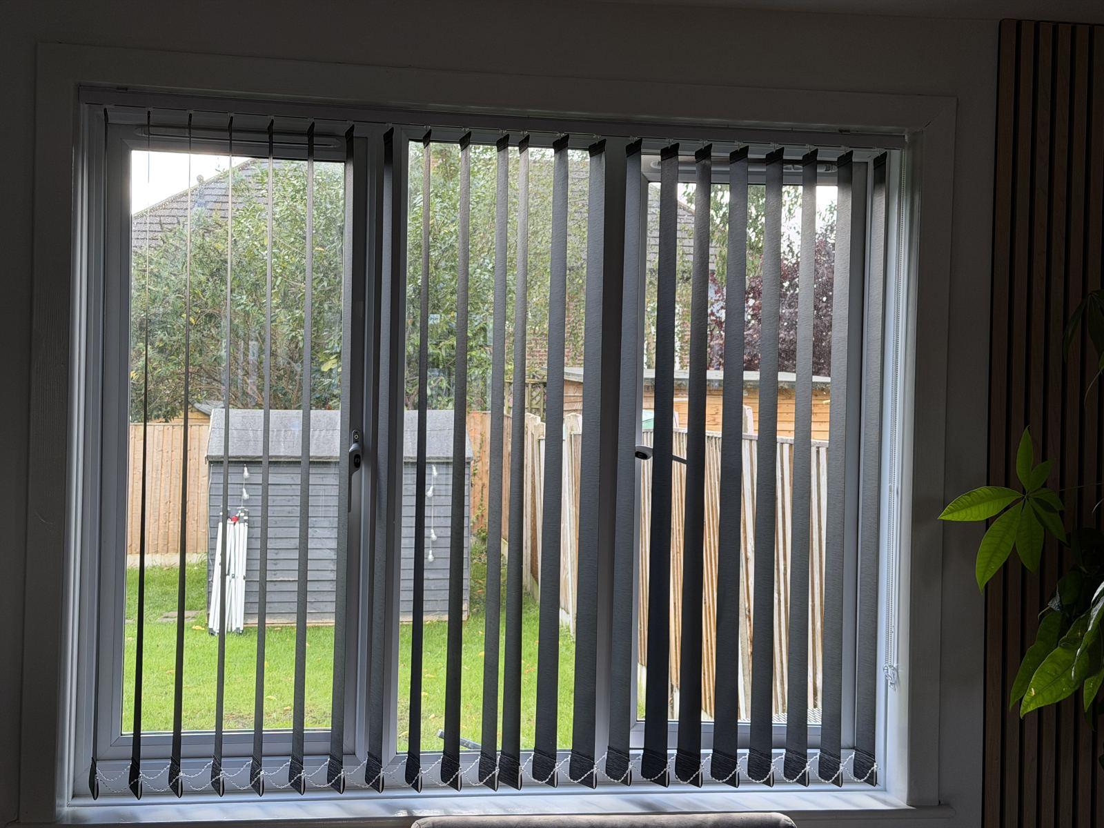 Grey vertical blinds fitted across a wide double window looking onto a garden